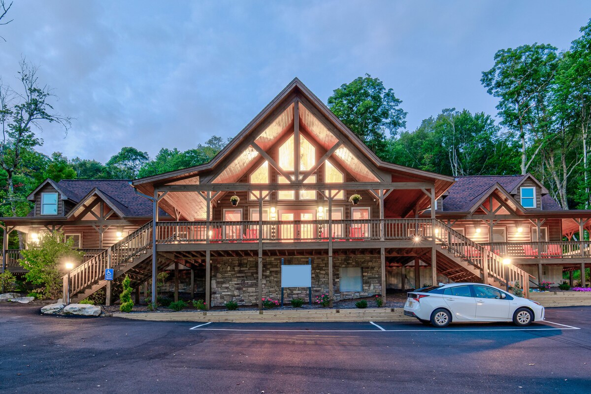 The exterior of a spacious lodge features a distinct A-frame design with large windows that illuminate the entrance. A wide staircase leads to the main entrance, surrounded by landscaped grounds. A parking area is visible, accommodating vehicles.