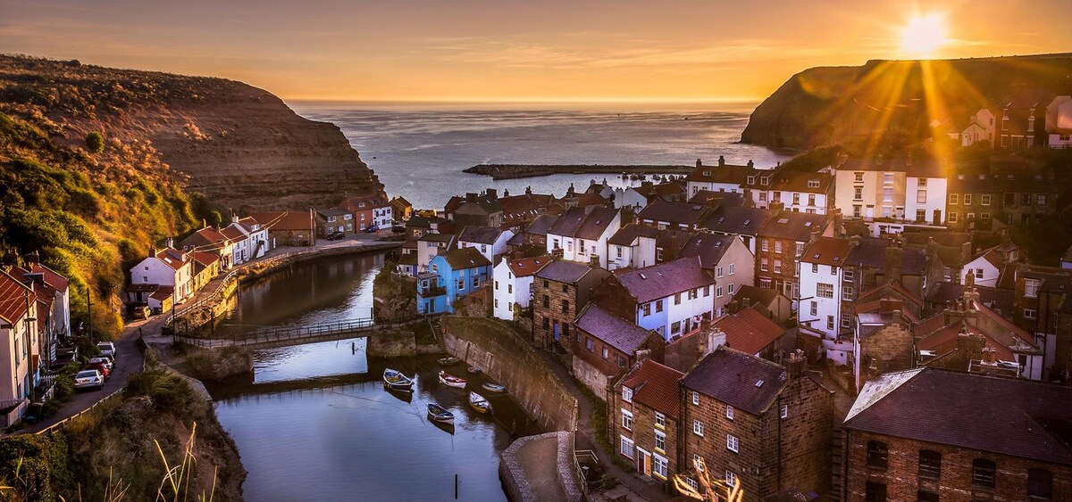 A tranquil coastal village is depicted at sunset, with houses lining the harbor that reflects the warm hues of the sky. A calm sea is visible in the background, while boats are moored near the water's edge, surrounded by rolling hills.