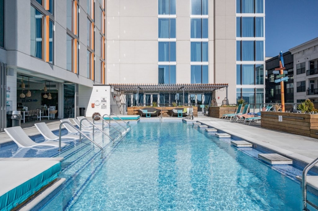 A large swimming pool is visible, featuring clear blue water and comfortably arranged lounge chairs beside it. A shaded area with seating is provided at one end. The building's modern facade, characterized by multiple large windows, reflects sunlight, enhancing the overall inviting outdoor space.