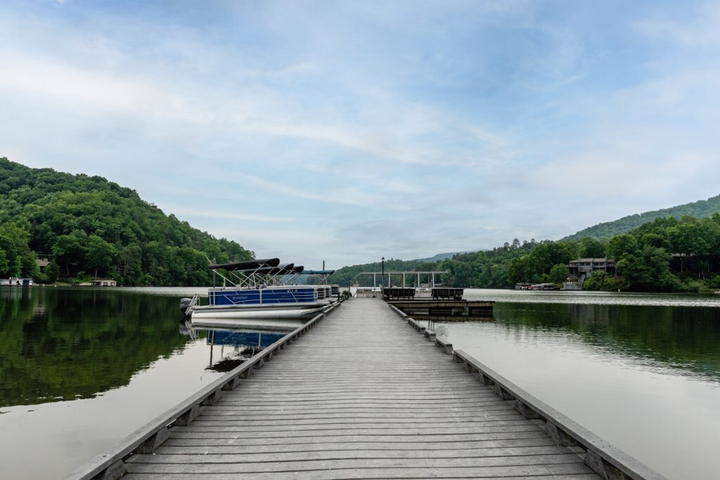 A wooden dock extends towards a calm lake, framed by lush green hills. Reflections of the surroundings shimmer on the tranquil water. A pontoon boat is visible at the dock's end, surrounded by the serene landscape.