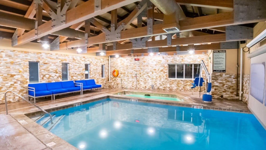 A well-lit indoor pool area is featured, with spacious seating along the side. The pool has clear water and steps leading in. The walls are decorated with stone accents, and large windows provide natural light. A safety buoy is displayed on the wall.
