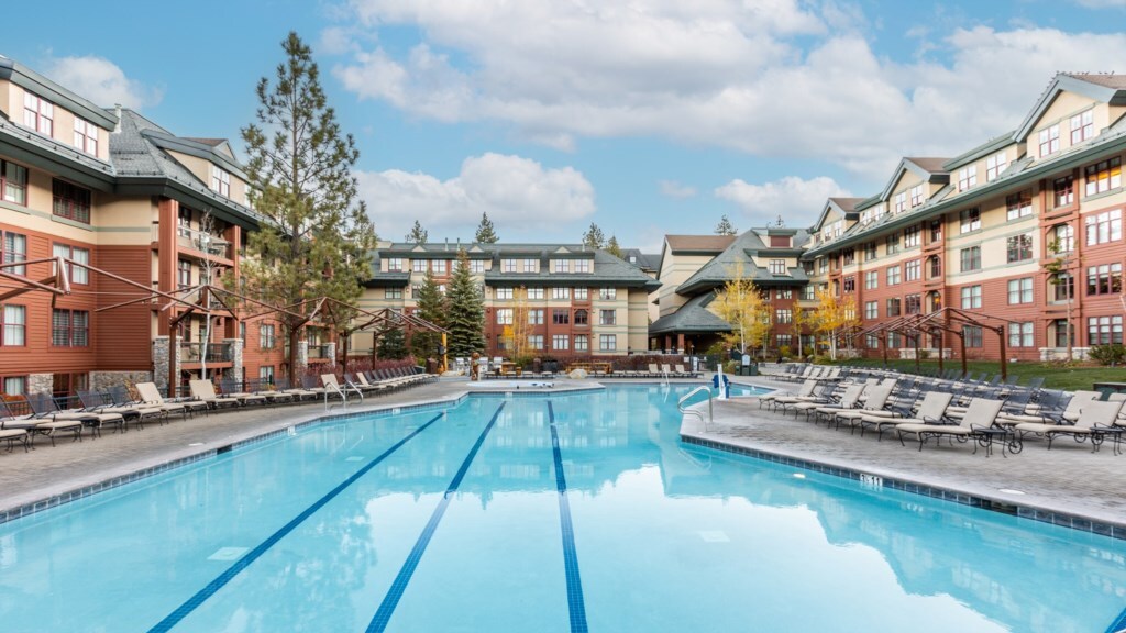 An outdoor pool is featured, reflecting the surrounding trees and buildings. Lounge chairs are arranged along the poolside, creating a spacious area for relaxation. The multi-story resort building showcases a combination of wood and stone textures, set against a clear blue sky.