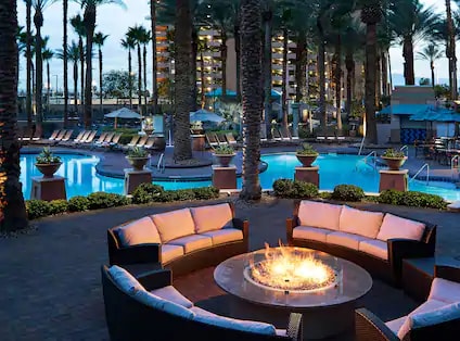 An inviting outdoor seating area features a circular arrangement of cushioned chairs around a fire pit. Tropical palm trees surround a tranquil pool area, where lounge chairs are positioned alongside shimmering water, creating a relaxed atmosphere for guests.