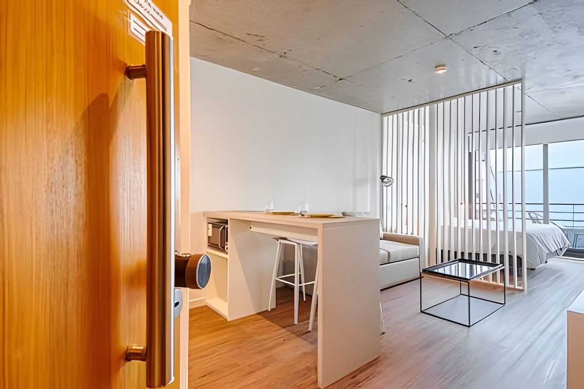 A modern entryway offers a glimpse into a minimalist interior. A small dining table with bar stools is visible alongside a sleek lounge area. A partition separates the cozy sleeping area, which features a spacious bed, while natural light flows in through large windows.