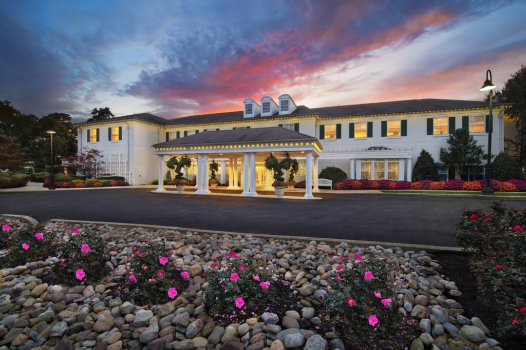The exterior of the resort features a grand entrance highlighted by a portico. Colorful flower beds with pink blooms surround the entryway. The building's facade is adorned with large windows and a classic architectural design, set against a vibrant sunset sky.