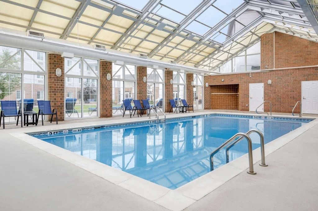 An indoor pool is featured with a clear roof allowing natural light to brighten the space. Surrounding the pool are several blue lounge chairs positioned for comfort. Large windows provide views to the outside, enhancing the serene atmosphere.