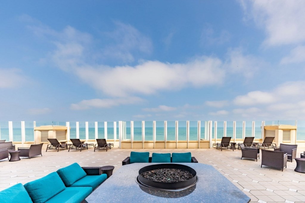 An outdoor lounge area features comfortable seating in shades of turquoise and dark brown. A circular fire pit sits at the center, surrounded by a smooth stone surface. In the background, unobstructed views of the ocean and blue sky create a serene atmosphere.