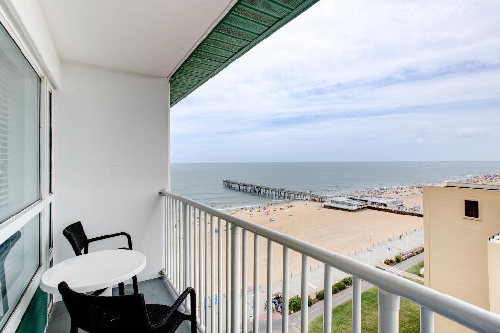A balcony offers a stunning view of the beach and the ocean. Two black chairs and a round table are positioned on the balcony, creating a space for relaxation. The expansive shoreline, dotted with beachgoers and umbrellas, stretches out towards the horizon.