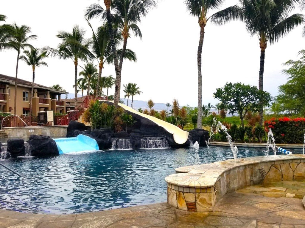A refreshing outdoor pool is surrounded by palm trees, showcasing a water slide and gentle waterfalls. The area is designed with natural stone accents and features a seating space along the pool's edge, inviting relaxation in a tropical setting.
