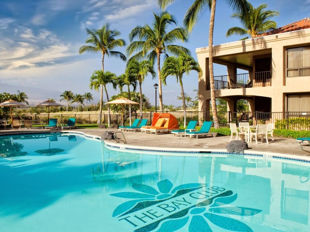 A spacious swimming pool is surrounded by vibrant green palm trees and comfortable lounge chairs. Colorful umbrellas provide shade near the poolside. The building in the background features a balcony and multiple windows, enhancing the tropical resort atmosphere.