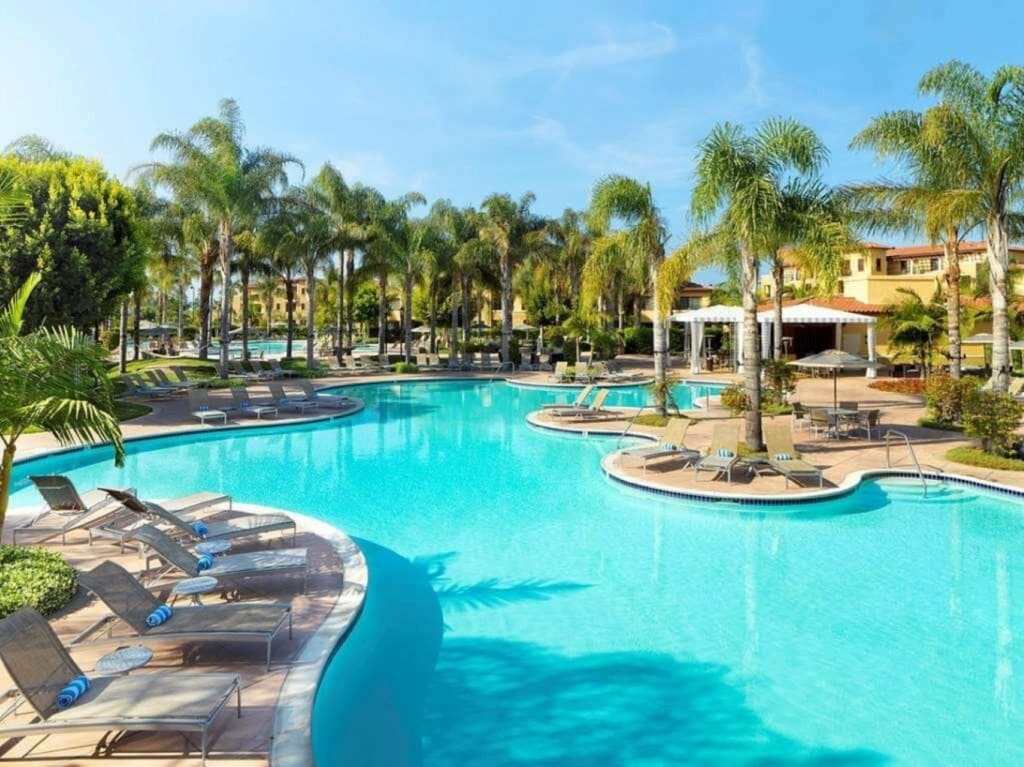 A large, inviting pool area is surrounded by palm trees and tropical landscaping. Lounge chairs are positioned along the water's edge, creating a relaxing atmosphere. A shaded patio area can be seen in the background, providing a space to unwind.