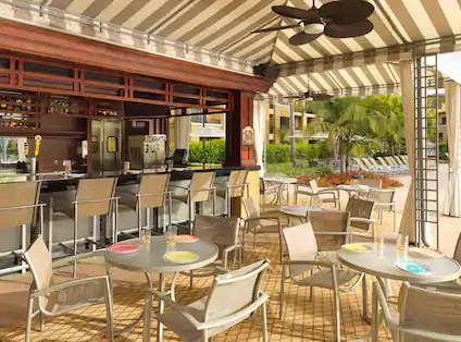 An outdoor seating area is set under a striped canopy, featuring several round tables with light-colored tops and metal chairs. A bar with a variety of beverages is situated in the background, surrounded by lush greenery and loungers visible in the distance.