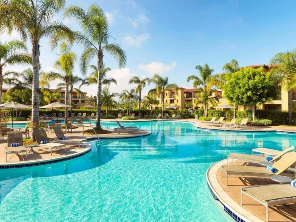 A spacious outdoor pool is surrounded by palm trees and loungers, providing ample seating. Shade umbrellas are positioned throughout the area, and the vibrant water reflects the bright blue sky. Nearby buildings exhibit Spanish Mediterranean architecture, enhancing the resort atmosphere.