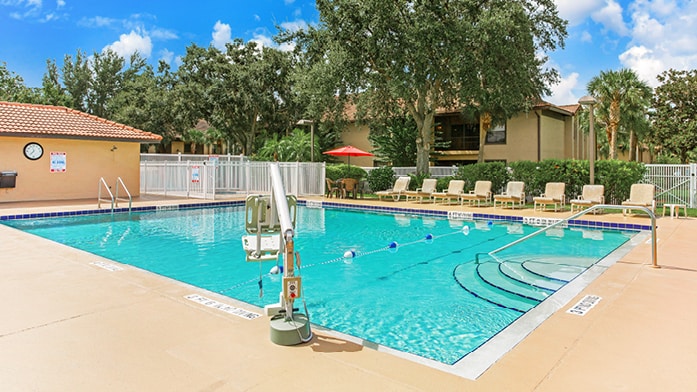 A large swimming pool is surrounded by lounge chairs, providing ample space for relaxation. A shallow entry area is visible, facilitating easy access. Umbrellas can be seen offering shade near the poolside. Lush greenery frames the area, contributing to a peaceful outdoor setting.