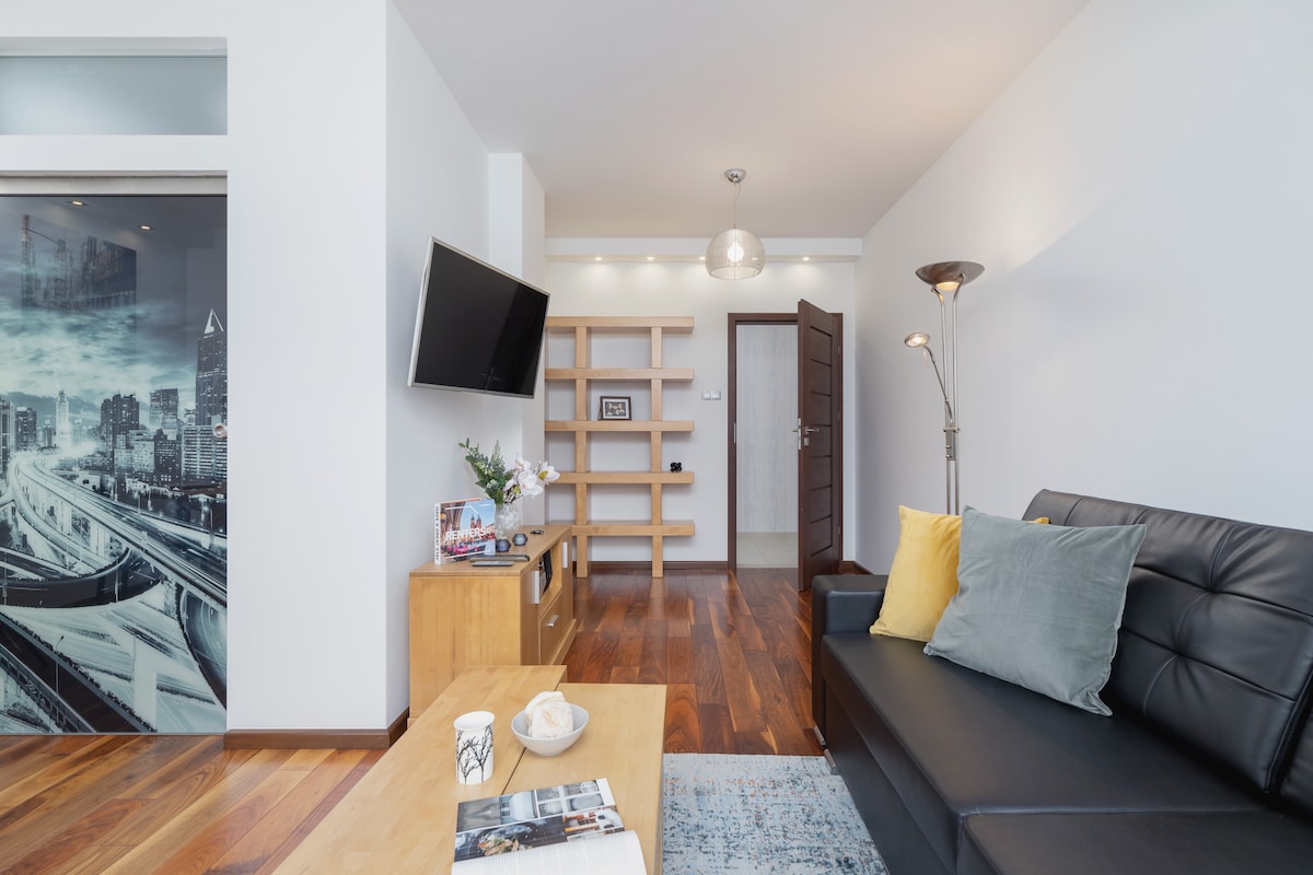 A bright living room features a black sofa with yellow and blue cushions, a wooden coffee table, and a flat screen TV mounted on the wall. Light wood accents and a modern shelving unit create a cozy atmosphere, while large windows allow natural light to fill the space.