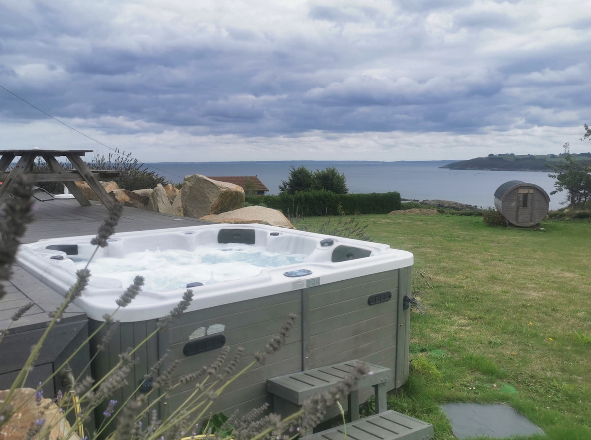 A jacuzzi overlooks the sea, surrounded by a neatly maintained lawn. Nearby, a wooden picnic table provides an inviting spot for outdoor meals. The scenic view includes rocky outcrops and a distant coastline, under a sky partly covered with clouds.
