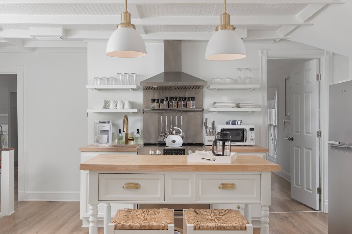 The modern kitchen features white cabinetry and an island with bar seating. Stainless steel appliances, including a stove and refrigerator, are visible. A sleek hood vent hangs above the stove, while open shelving displays dishware and spice containers.