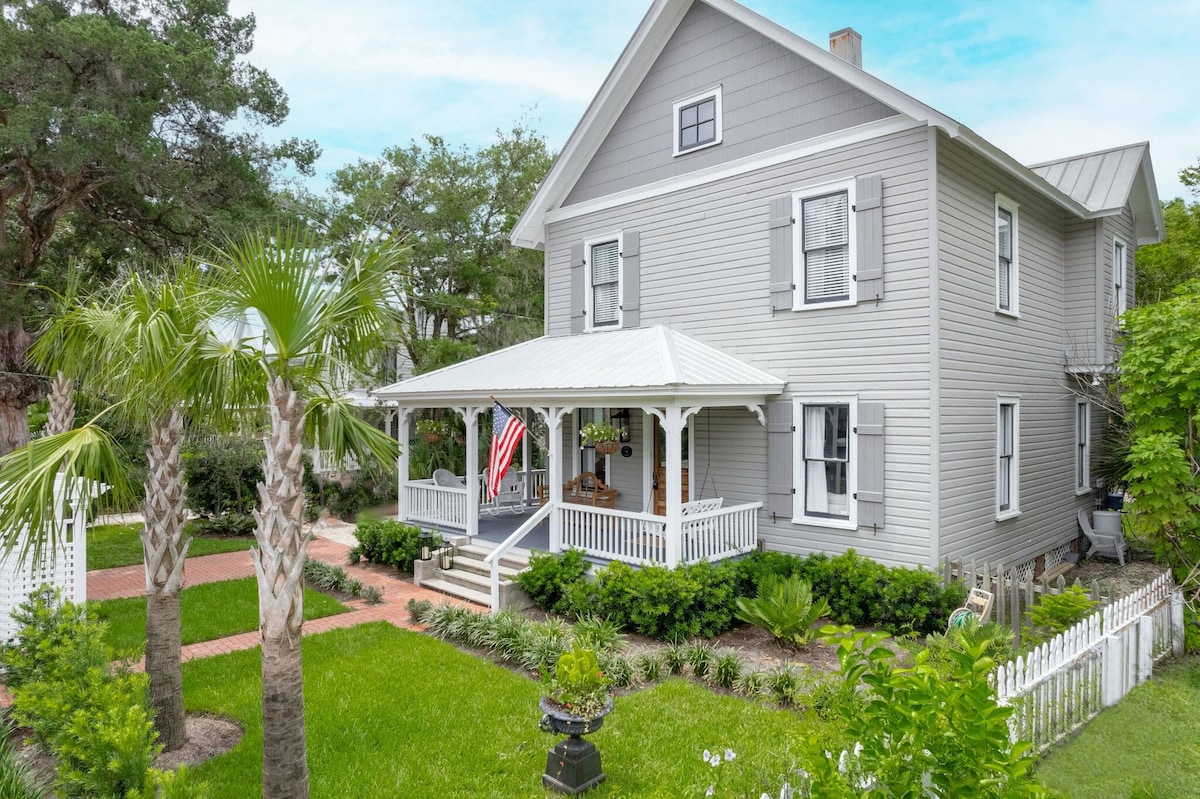 A charming historic home is featured, showcasing a wrap-around porch with a gabled roof and white railings. Lush greenery surrounds the property, complemented by palm trees and well-maintained landscaping. The exterior is painted in soft grey, enhancing its classic appeal.