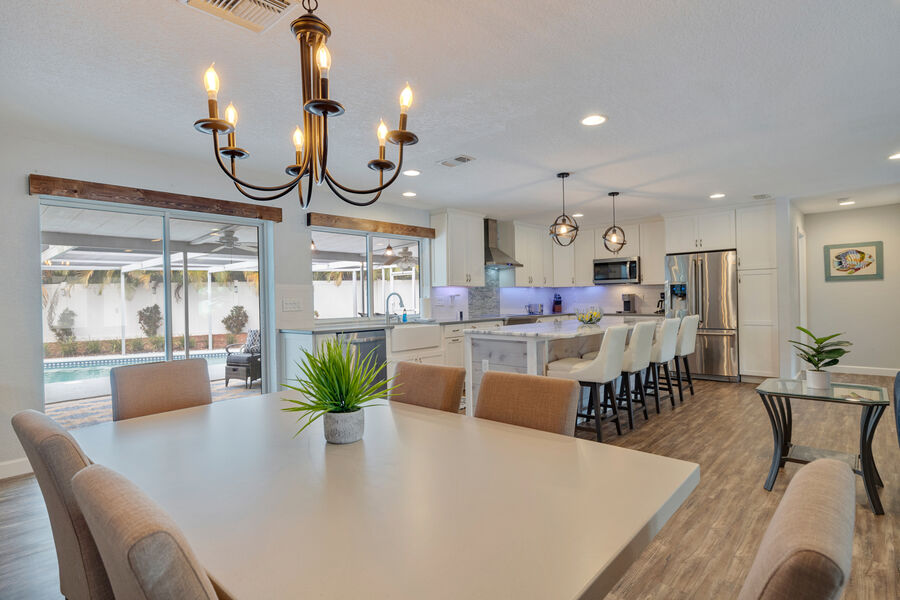 An open-concept kitchen and dining area with a large dining table surrounded by several upholstered chairs. A modern chandelier illuminates the space, which features a kitchen island with seating for four and stainless steel appliances in the background.
