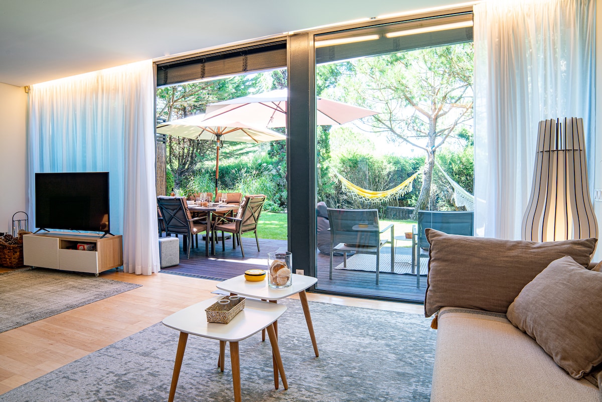 A living area features large glass doors that open to a garden patio. A cozy sofa and textured area rug are visible, while a stylish TV unit provides entertainment. Outdoor furniture and an umbrella can be seen through the doors, along with a hammock hanging in the greenery.