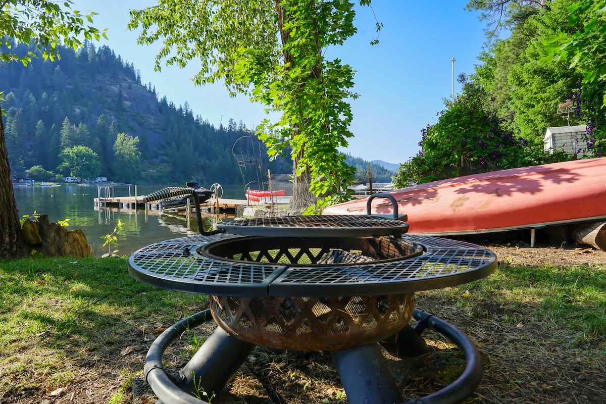 A fire pit is positioned near the lake, surrounded by vibrant green grass and bordered by lush trees. The calm water reflects the surrounding hills, with kayaks and docks visible in the background, creating a serene lakeside setting.