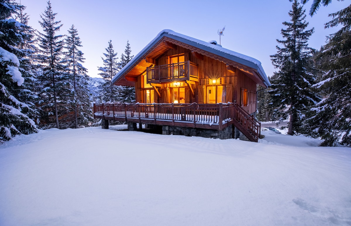 The chalet is set amidst a snowy landscape, showcasing its wooden exterior and warm, glowing lights from large windows. The expansive deck wraps around the house, bordered by tall trees that enhance the feeling of seclusion and tranquility.