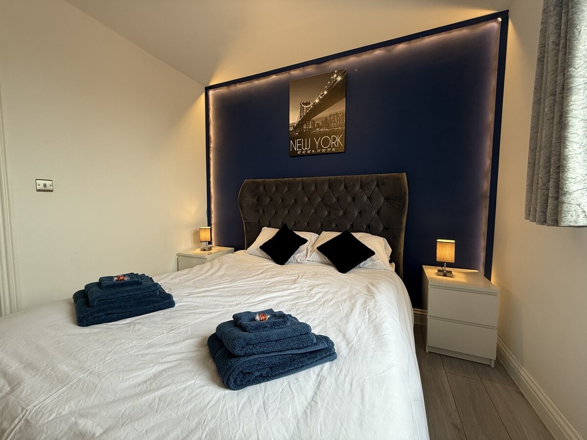 A modern bedroom features a spacious bed with a tufted headboard against a deep blue accent wall. Nightstands with lamps are positioned on either side, and neatly folded towels are arranged on the bed. Natural light filters through a window with light drapes.