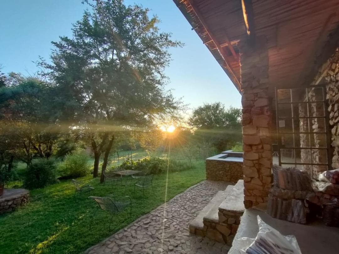 The image captures a warm sunset casting golden light onto a stone patio. A stone structure is visible, with a glimpse of a splash pool at the edge. Several chairs are positioned in the grassy area, surrounded by trees and greenery.