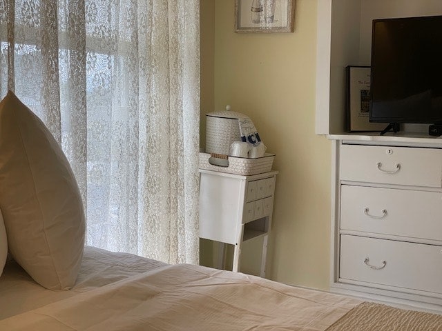 A cozy bedroom corner features a spacious bed with two large pillows. A white lace curtain gently filters light from the window, adding a soft glow. A small white nightstand sits beside the bed, adorned with a decorative piece. A flat-panel TV is positioned on a nearby dresser.