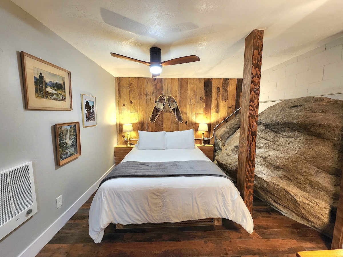 A serene bedroom features a queen bed with two bedside lamps on wooden nightstands. Artwork adorns the walls, and natural wood panels serve as a backdrop, complemented by a unique granite rock accent. A ceiling fan provides cooling in this cozy and inviting space.