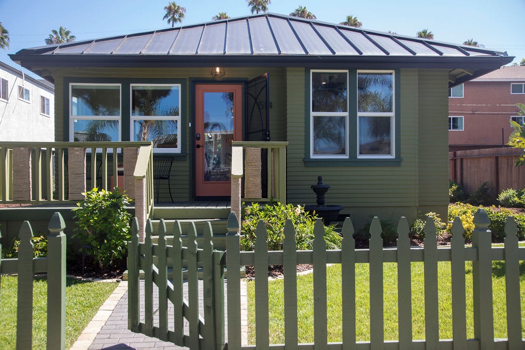 The front view of a charming green house features a welcoming porch with a metal roof and a fenced yard. Lush landscaping frames the property, complemented by palm trees in the background. A vibrant front door adds a splash of color, inviting guests to enter.