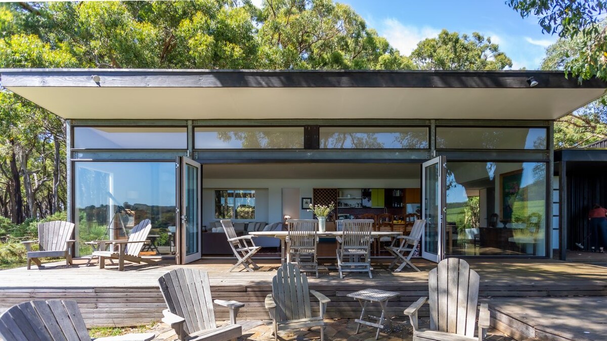 A modern house features large glass sliding doors that open to a spacious deck. Outdoor seating arrangements include wooden chairs, creating an inviting space for relaxation. Lush greenery surrounds the area, enhancing the connection with nature.
