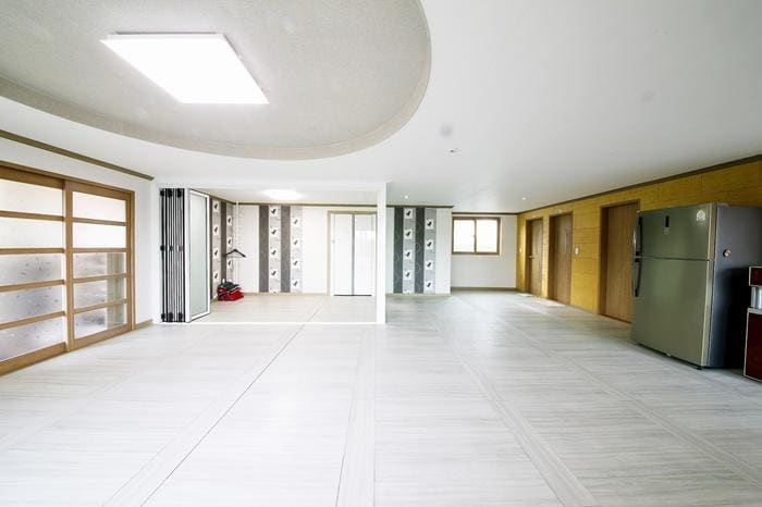A spacious living area features large, bright windows, allowing natural light to fill the space. The floor is covered with light-colored tiles, and sliding glass doors lead to another room. A modern refrigerator is visible, complementing the well-maintained interior.
