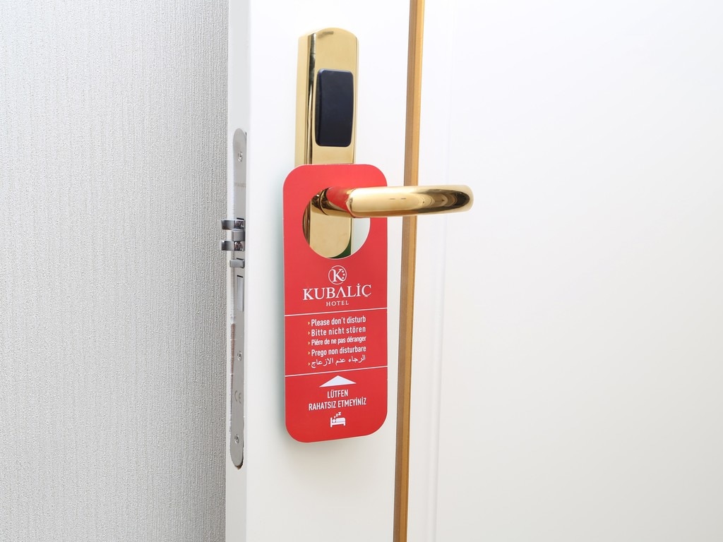A door equipped with a gold handle features a red do-not-disturb sign emblazoned with the Kubaliç Hotel logo. The clean, modern design of the door is complemented by subtle wall textures in the background.