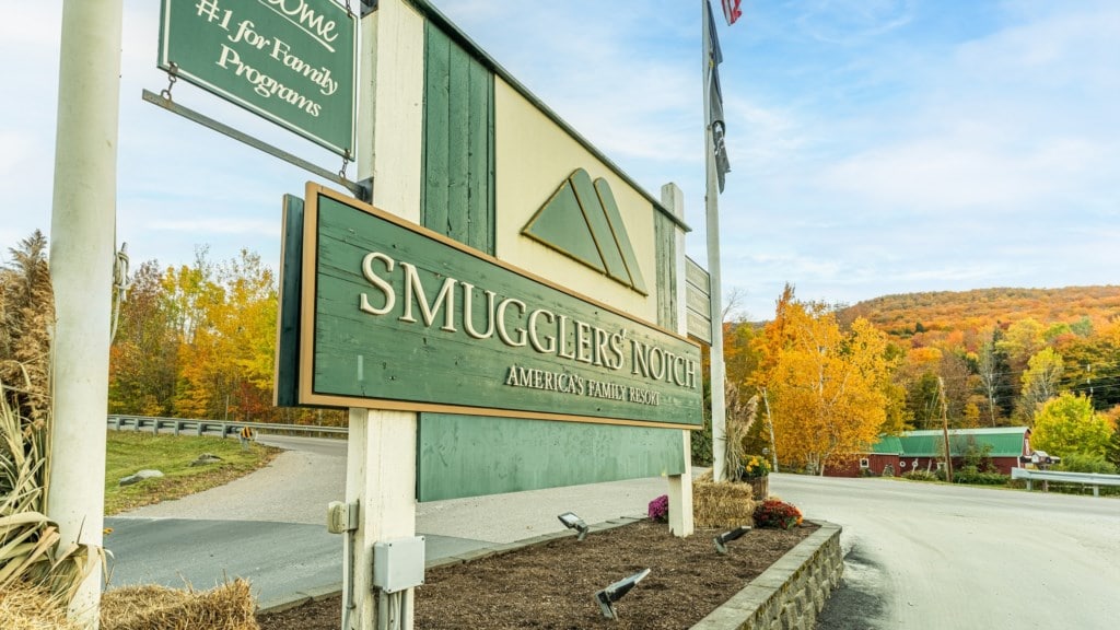 The entrance sign to Smugglers' Notch is prominently displayed, framed by colorful autumn foliage. The lush, green hills in the background complement the serene atmosphere of the resort village, while well-maintained shrubs and seasonal decorations enhance the welcoming entrance.