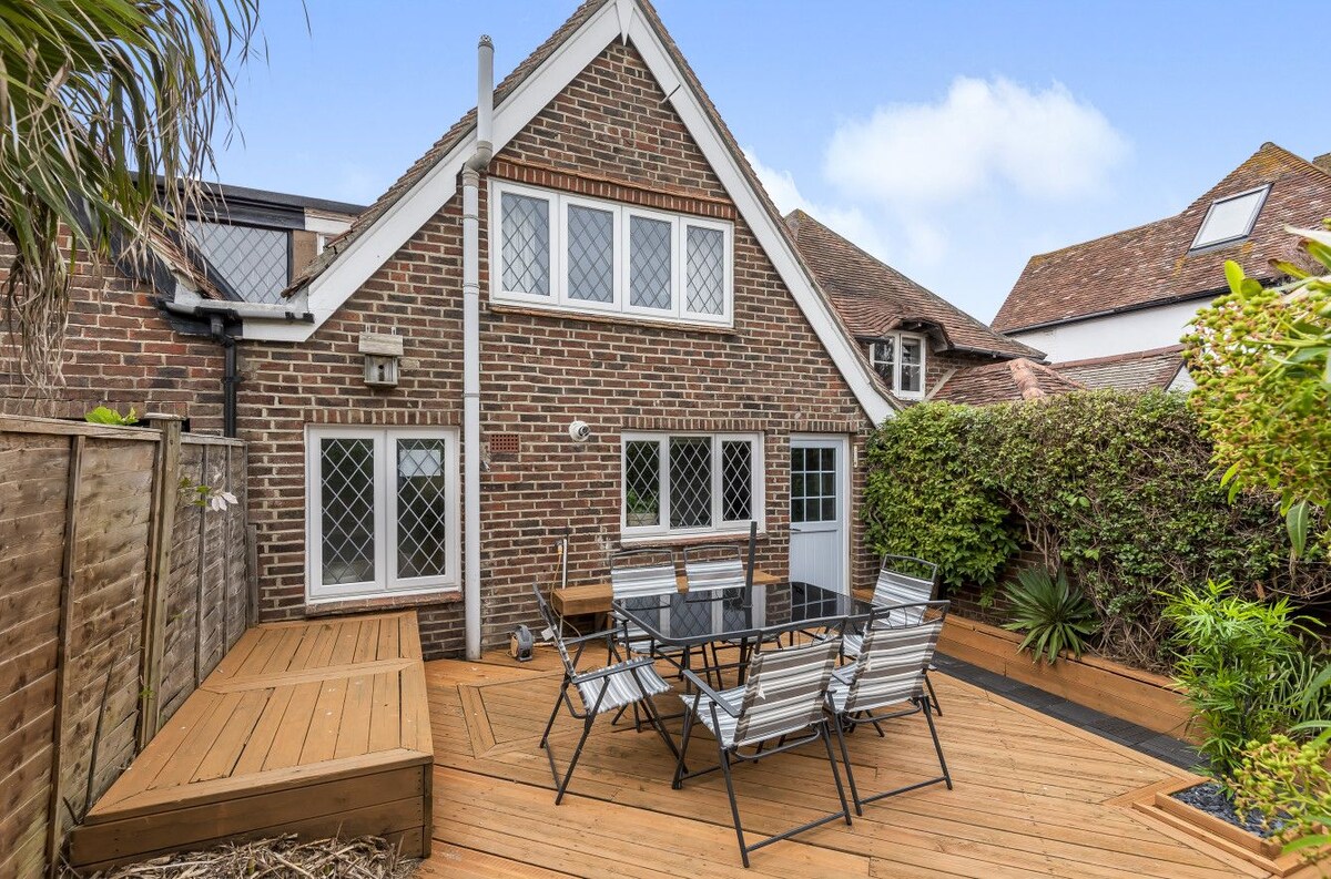 A small decked garden is visible, featuring a dining table surrounded by six chairs. The brick exterior of the cottage provides a charming backdrop, with greenery along the perimeter. Large windows allow natural light to enhance the outdoor space, creating a relaxed setting.