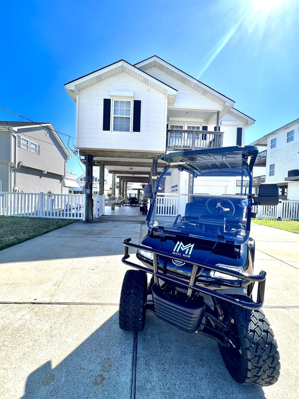 A modern golf cart is positioned in the foreground, with the raised house in the background. The home features a classic white exterior with black shutters, elevated on stilts above a concrete driveway. Sunlight reflects brightly off the scene, highlighting the outdoor space.