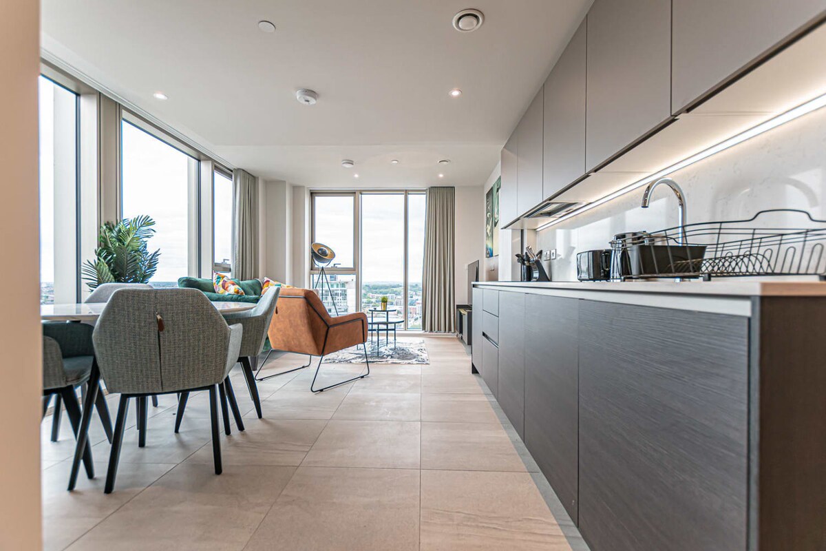 The open-plan living and dining area features modern furnishings in a neutral palette. A spacious kitchen with sleek cabinetry is visible on the left, while a dining table with upholstered chairs and a cozy seating area can be seen in the background, framed by large windows offering city views.