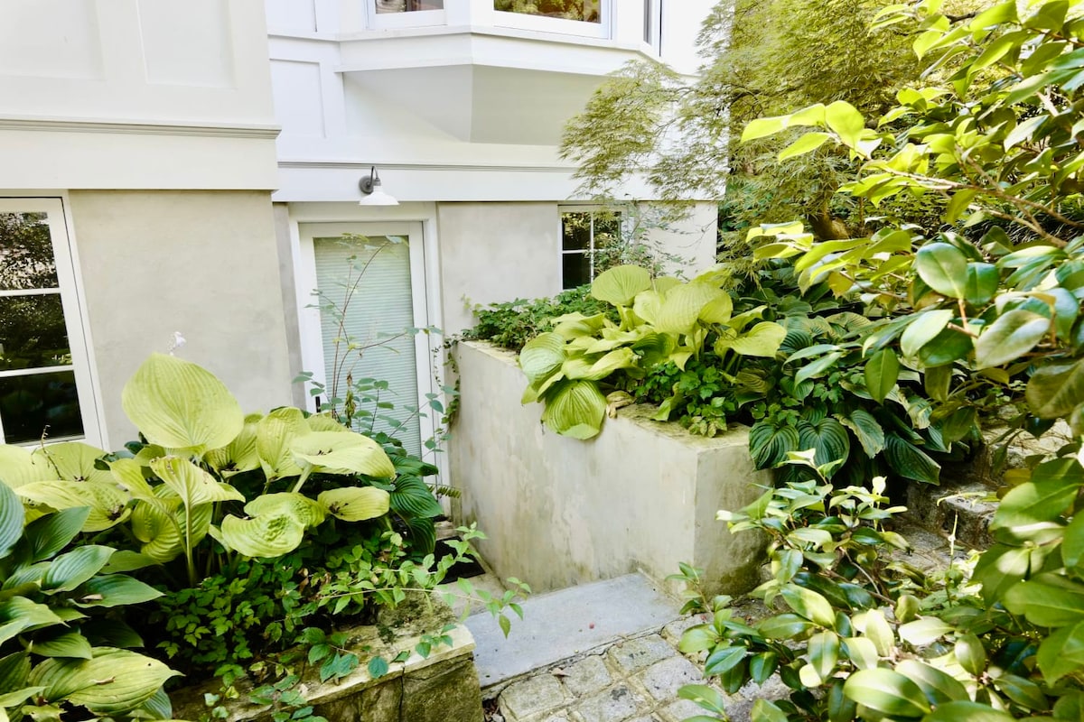 The private entrance to the apartment is framed by lush greenery, featuring various plants and foliage lining a narrow stone path. A discreet door is visible at the end of the pathway, providing access to the serene garden-level studio.