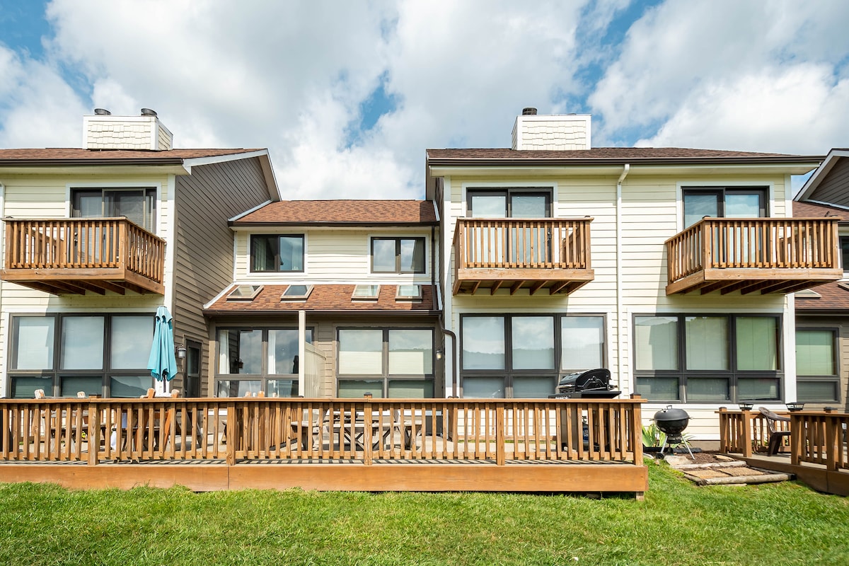 The exterior of the townhome showcases a two-story design with multiple balconies. Wooden decking extends across the front, leading to a grassy area. The structure is flanked by similar homes, and large windows are visible, allowing natural light into the living spaces.