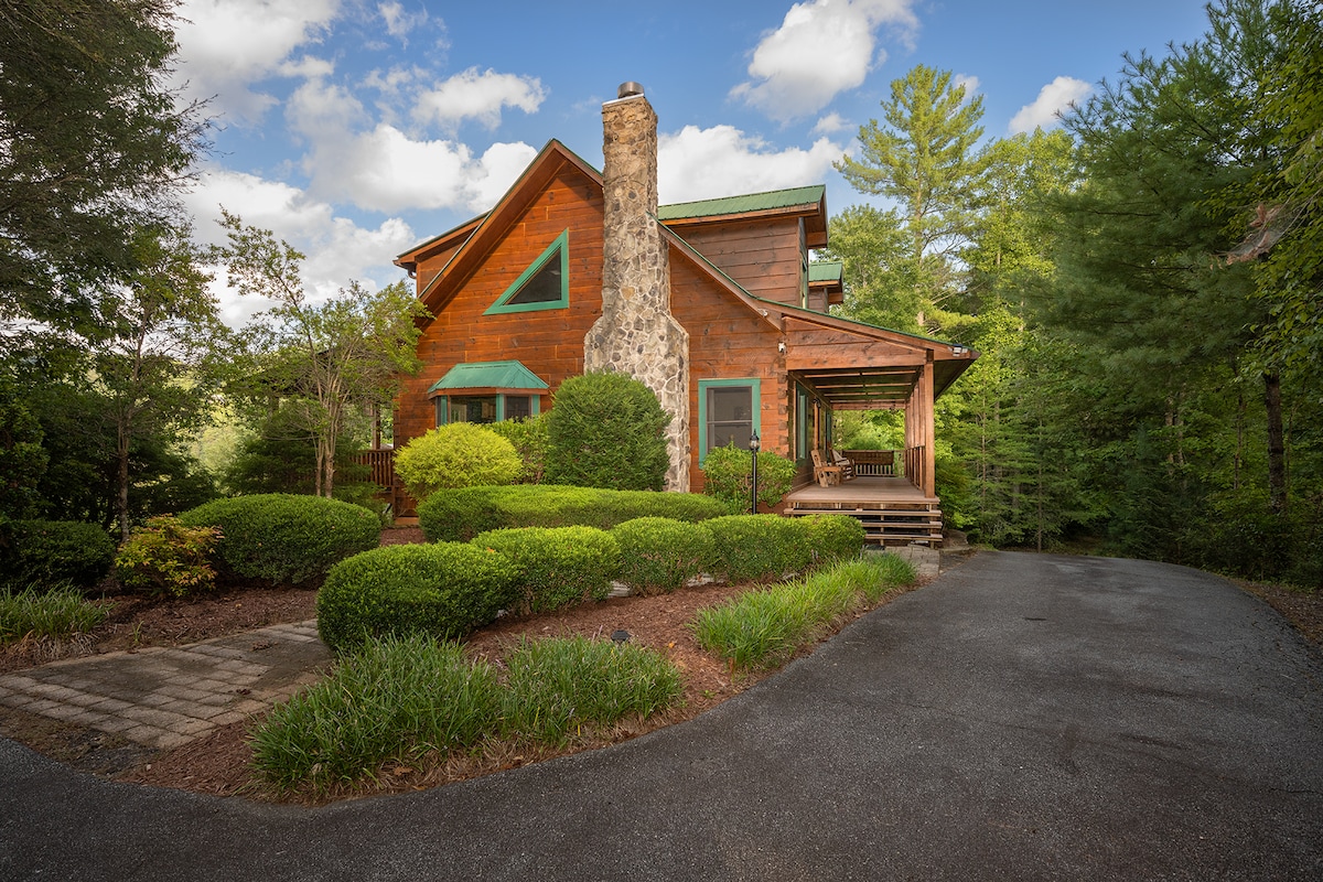 A charming mountain cabin is set against a backdrop of lush greenery. The wooden structure showcases a prominent stone chimney and is complemented by manicured shrubs and a warm inviting driveway leading to the entrance.