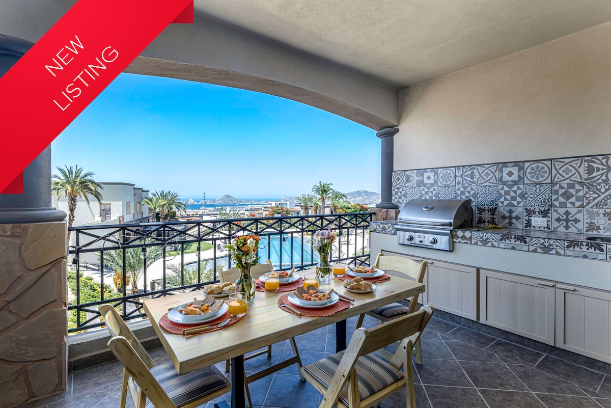 An outdoor dining area is showcased, featuring a table set with plates and drinks. A barbecue grill is visible nearby, with ocean views and palm trees in the background. The vibrant tiles on the wall add a decorative touch to the space.