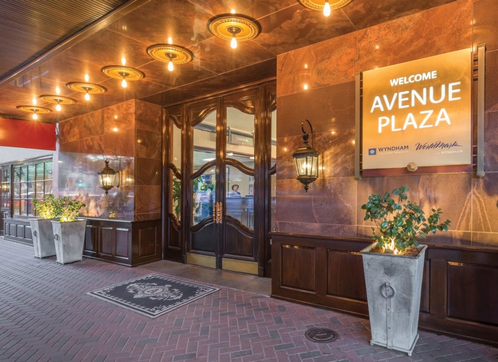 An elegant entrance showcases large wooden doors framed by polished stone walls. Warm lighting from vintage-style fixtures creates a welcoming glow. Potted plants with greenery are positioned beside the entrance, adding a touch of nature to the sophisticated exterior.