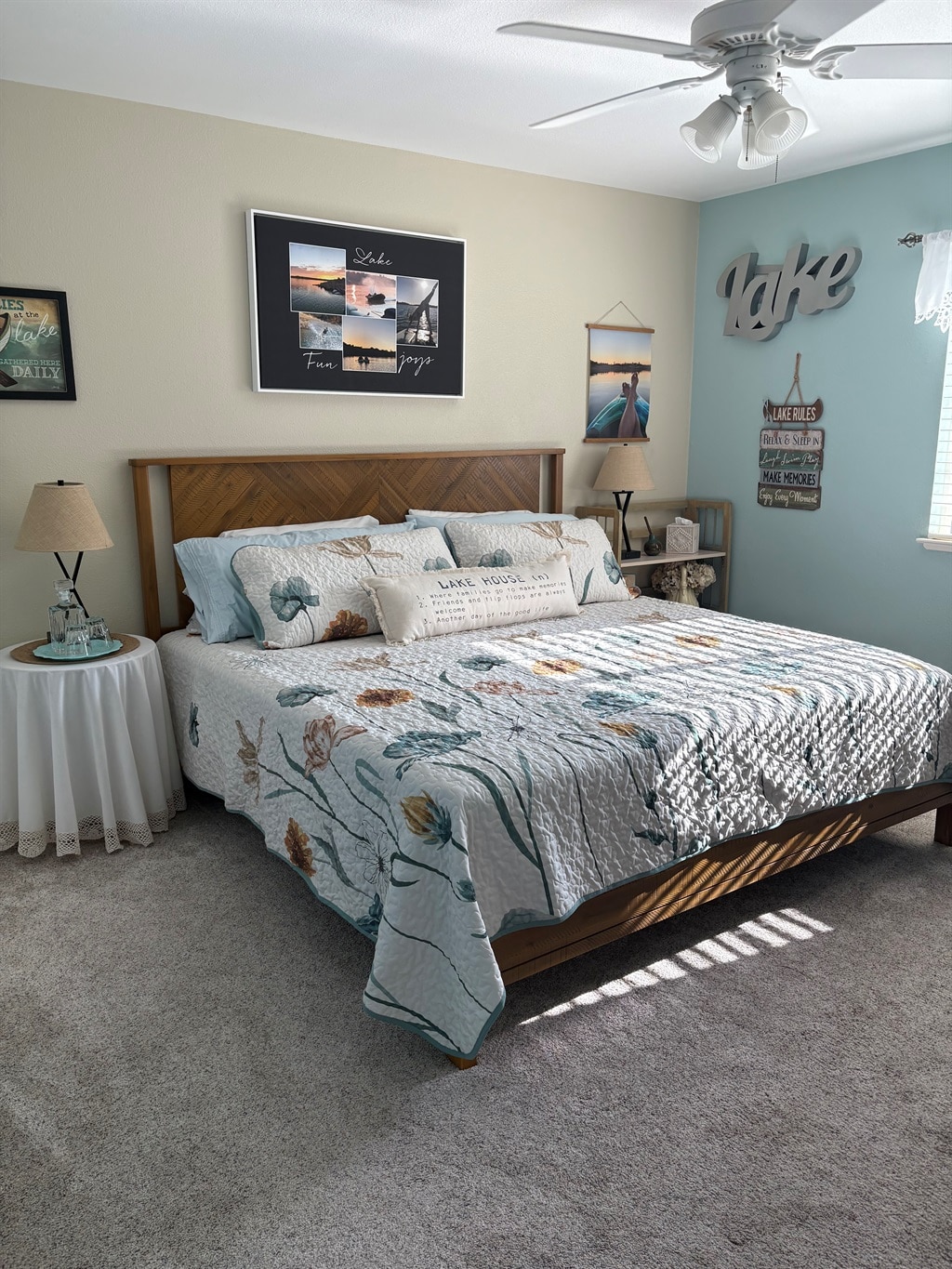 A comfortable bedroom features a wooden bed adorned with a decorative quilt showcasing lake-themed designs. Light blue walls complement the decor, and artwork is displayed above the bed. A bedside lamp is set on a small white table, and natural light filters through a window covered by light blinds.