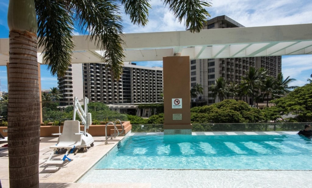 An outdoor pool area is viewed, featuring clear water and a modern design. Tall palm trees frame the scene, while a lounge chair is positioned nearby. The backdrop includes a large building and lush greenery, creating a relaxed atmosphere.