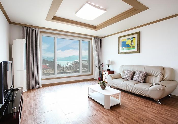 A bright living area features a comfortable sofa set against a large window, offering views of the sea. Natural light fills the space, highlighting the wooden flooring and minimalistic decor. A coffee table and decorative items complete the inviting atmosphere.