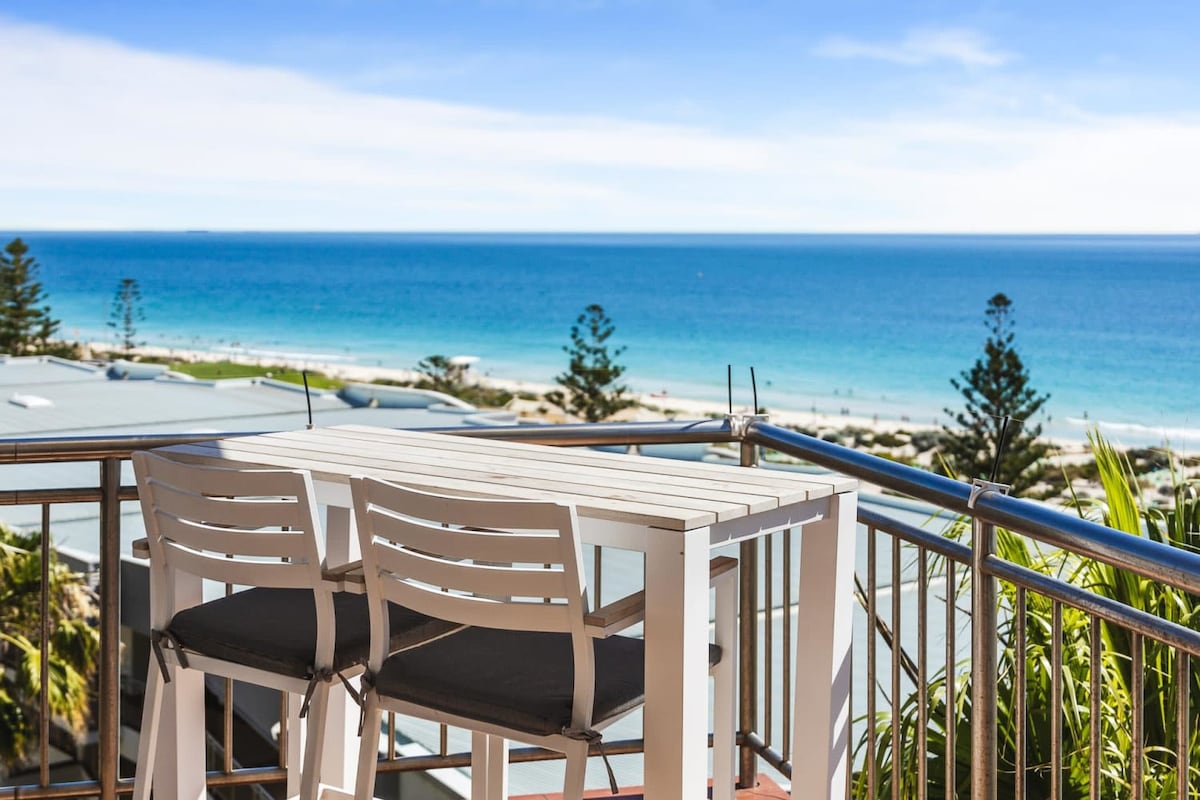 A scenic balcony view features a compact outdoor dining set with two chairs and a small table. The vast ocean stretches into the horizon, complemented by a clear blue sky and the sandy beach below, creating a tranquil seaside atmosphere.
