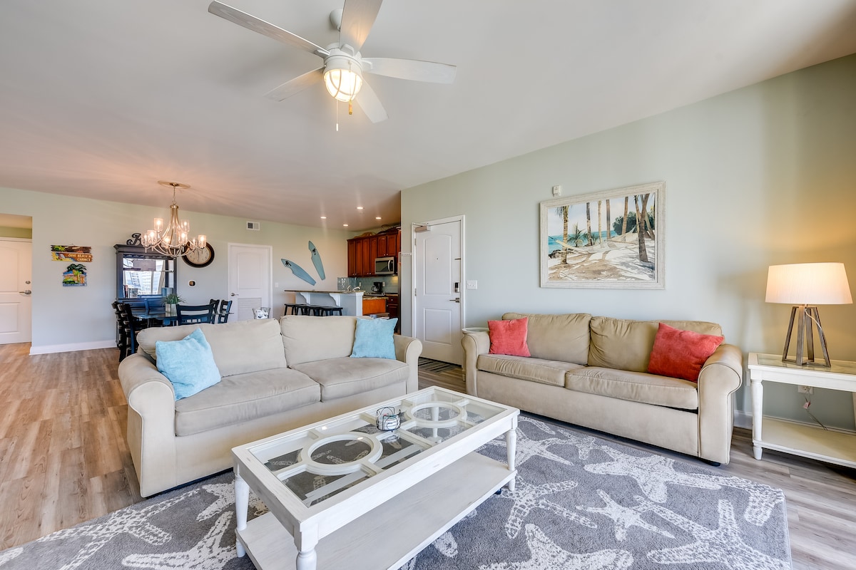 The living room features a light-filled space with two comfortable sofas, one showcasing vibrant coral cushions. A decorative coffee table sits in front, resting on a patterned area rug. A ceiling fan enhances airflow, while a large painting adorns the wall, offering a glimpse of coastal serenity.