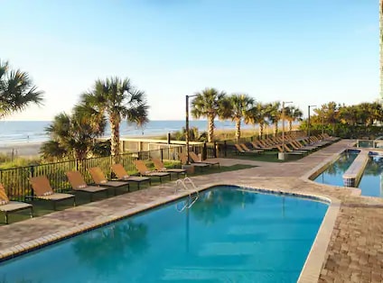 An outdoor pool area is surrounded by lounge chairs, offering relaxing views of the ocean. Lush palm trees frame the space, enhancing the coastal ambiance. A second pool can be seen nearby, inviting guests to enjoy the sun and refreshing water.