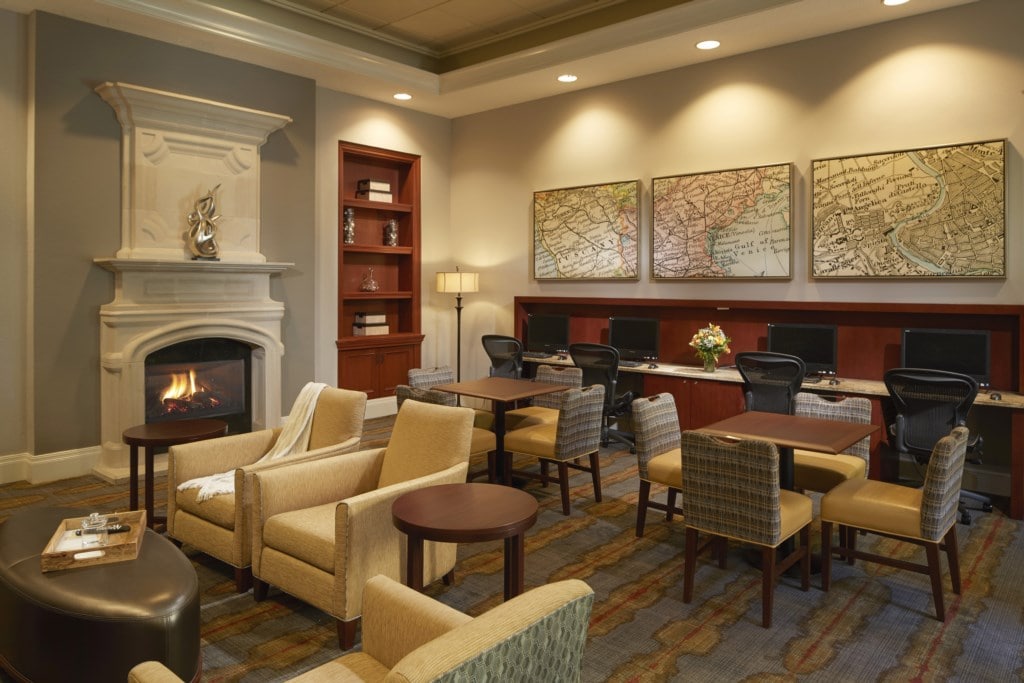 A comfortable lounge area is presented with soft seating arranged around small tables. A decorative fireplace is situated against the wall, while two maps are framed and hung above a wooden desk with computers. Warm light from a lamp adds to the welcoming ambiance.